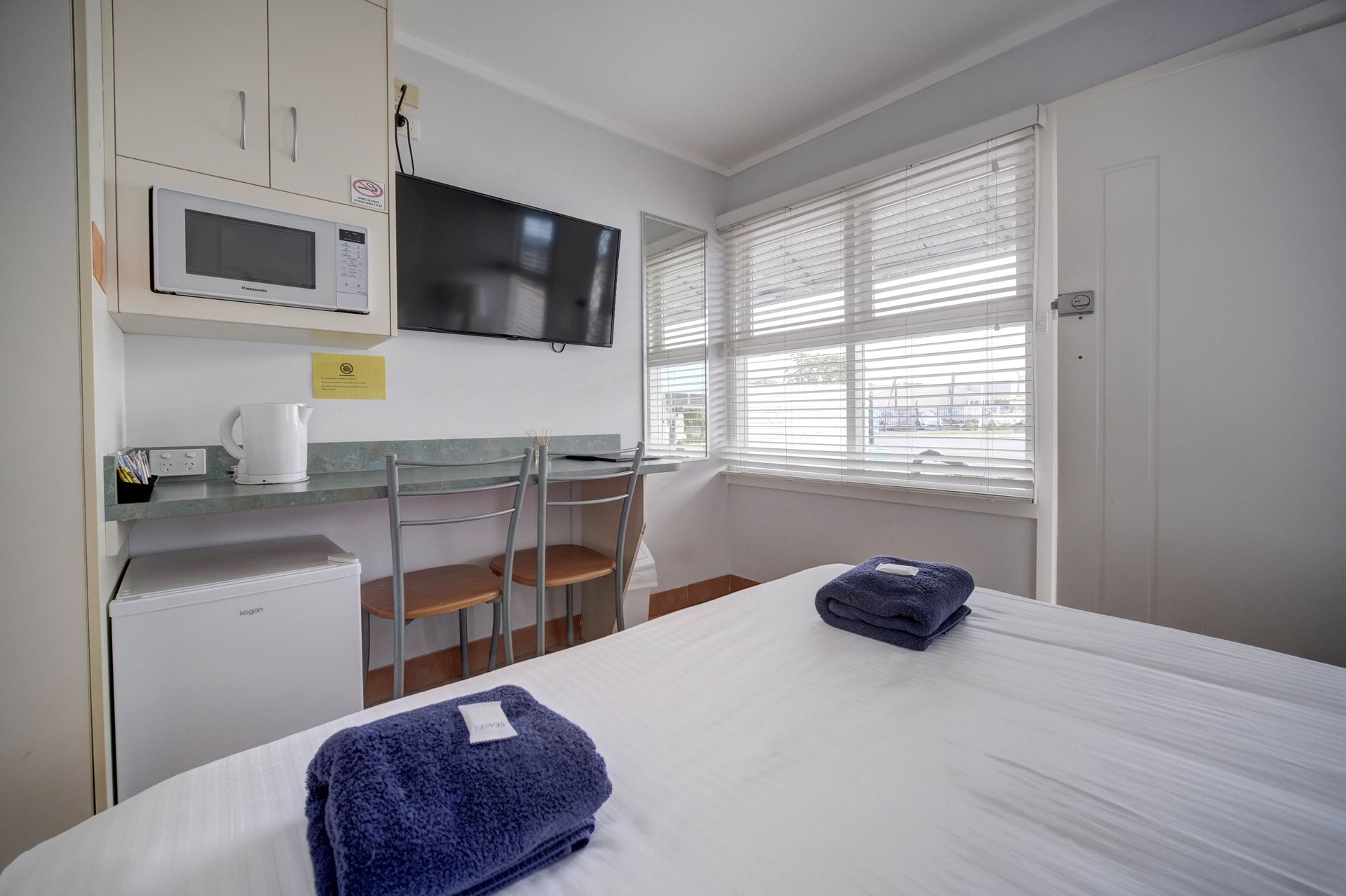 Small motel room with bed, TV, microwave, desk, and window with blinds. Two blue towels on the bed.