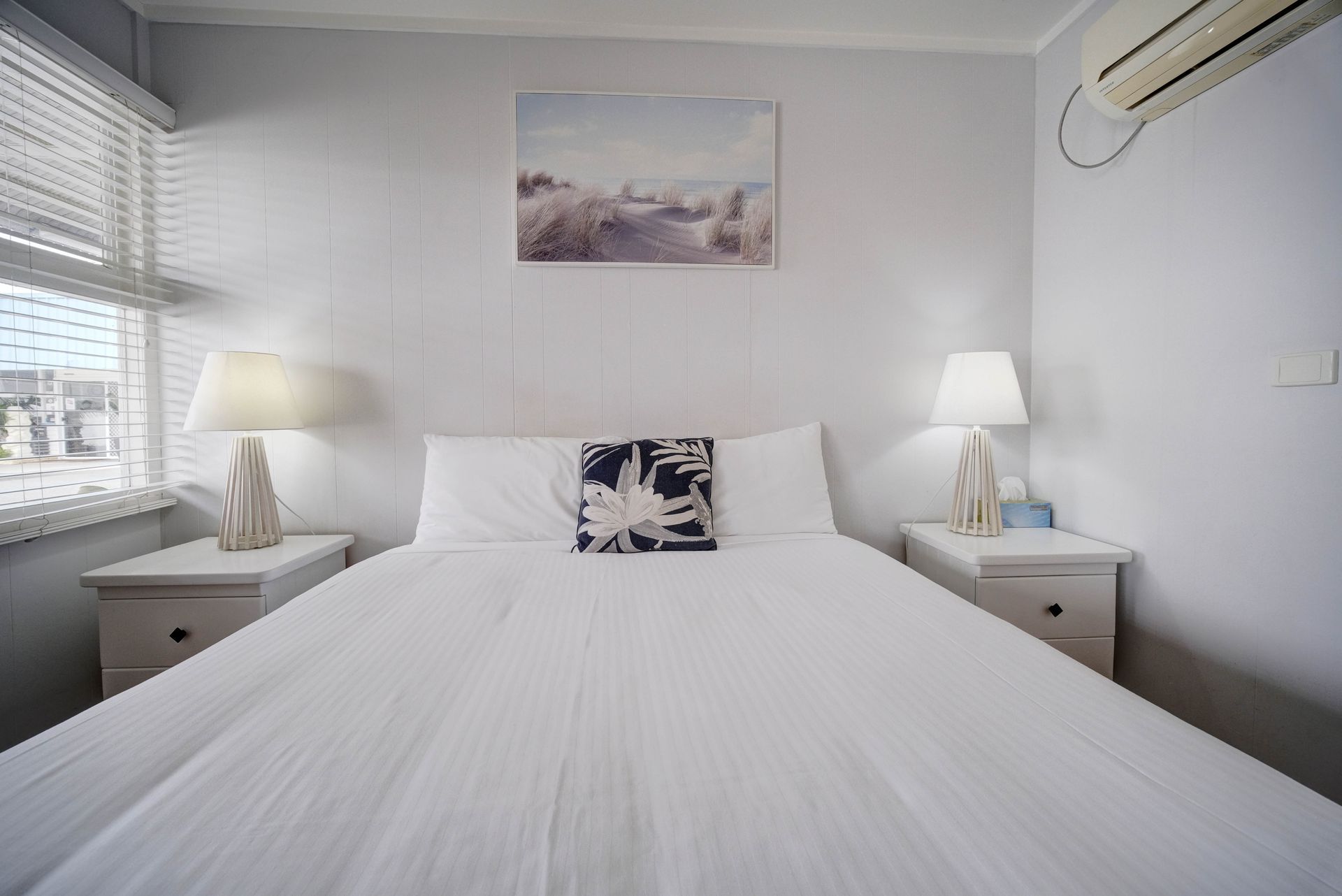 Bedroom with a white bed, lamps on nightstands, coastal artwork, and a window with blinds.