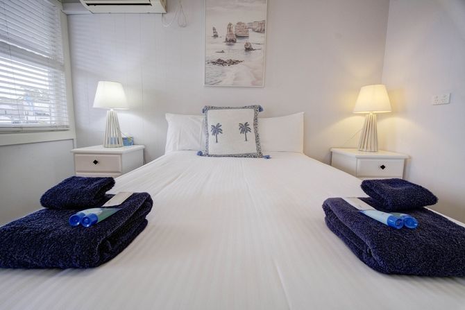 Bedroom with white walls, a bed with white linens, two lamps, and blue towels.
