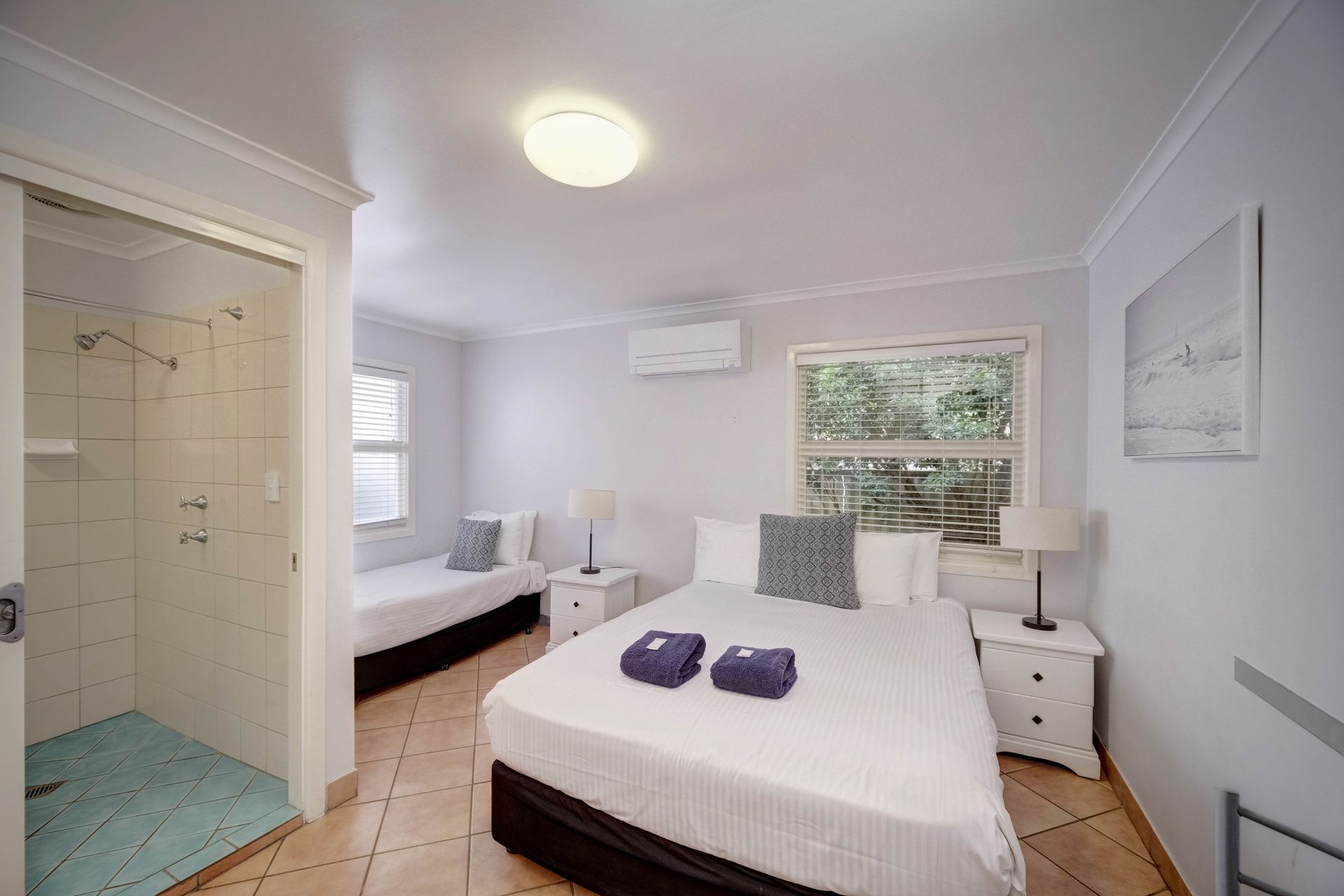 Bedroom with queen bed, single bed, shower, and windows. White walls, light wood floors.