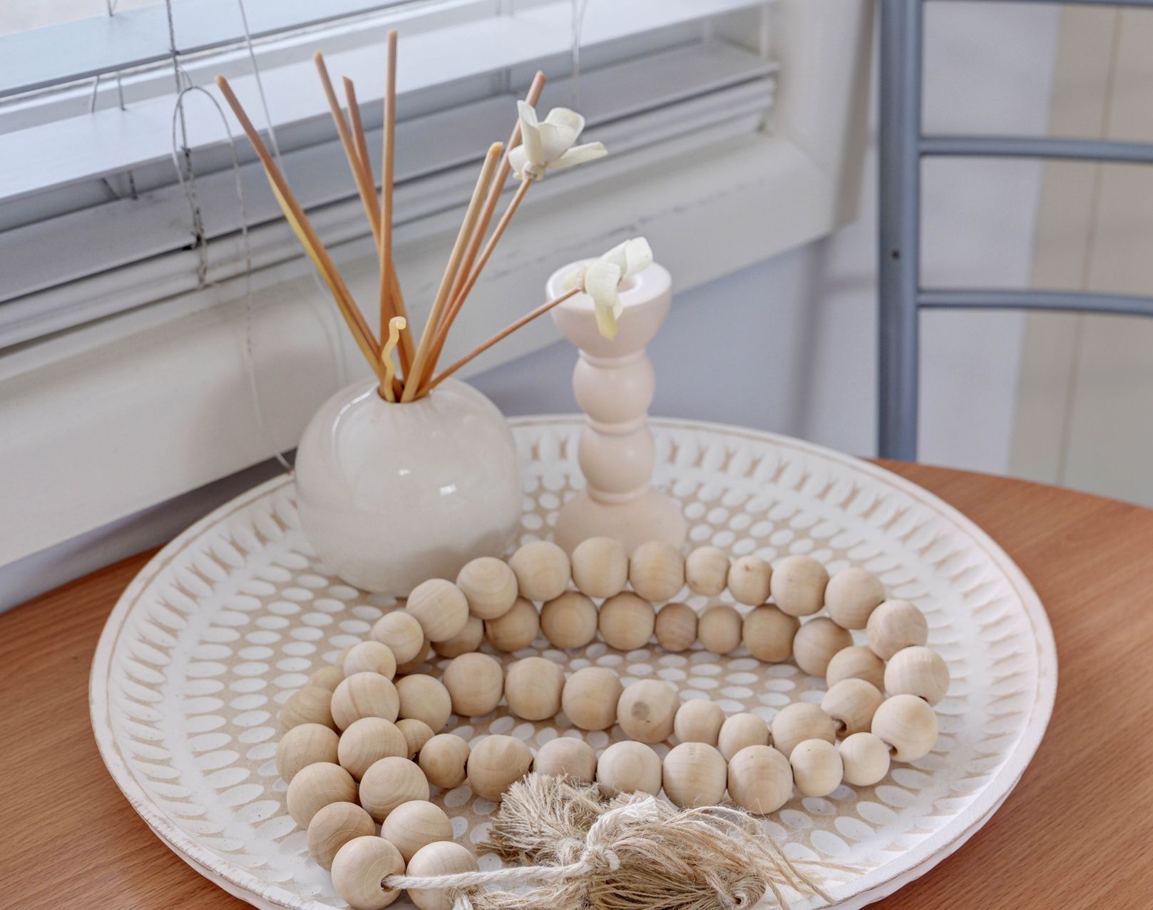 White decorative tray with wooden beads, diffuser, and candle holder near a window.
