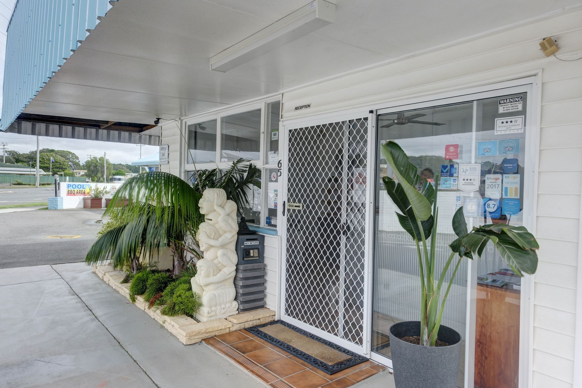Exterior of a small white shop with a glass door, potted plants, and a light-colored awning.
