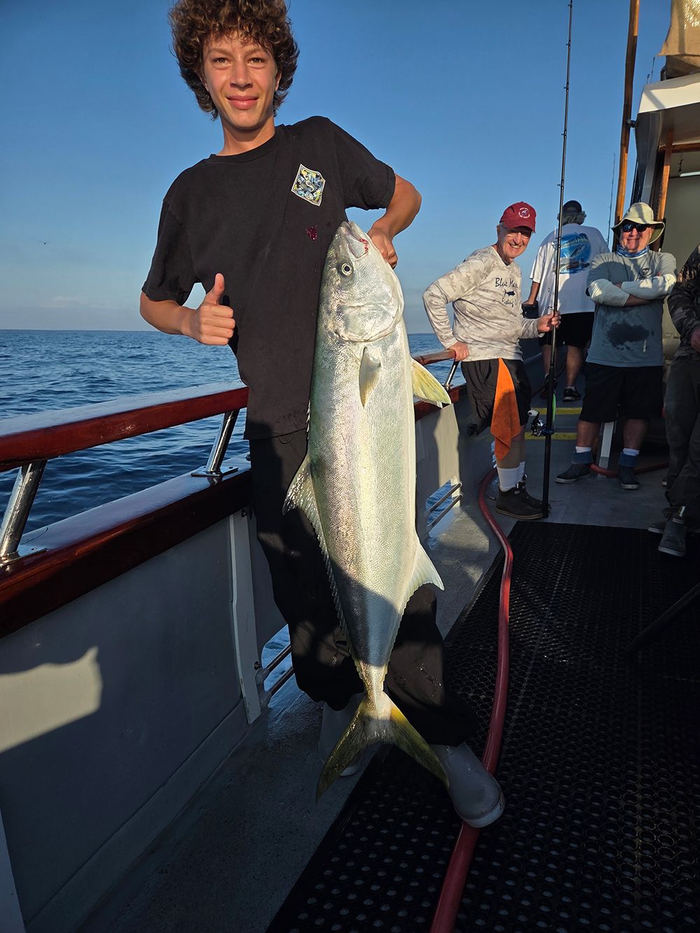 Boy holding a large yellowtail fish on a boat, giving a thumbs up. Ocean background.