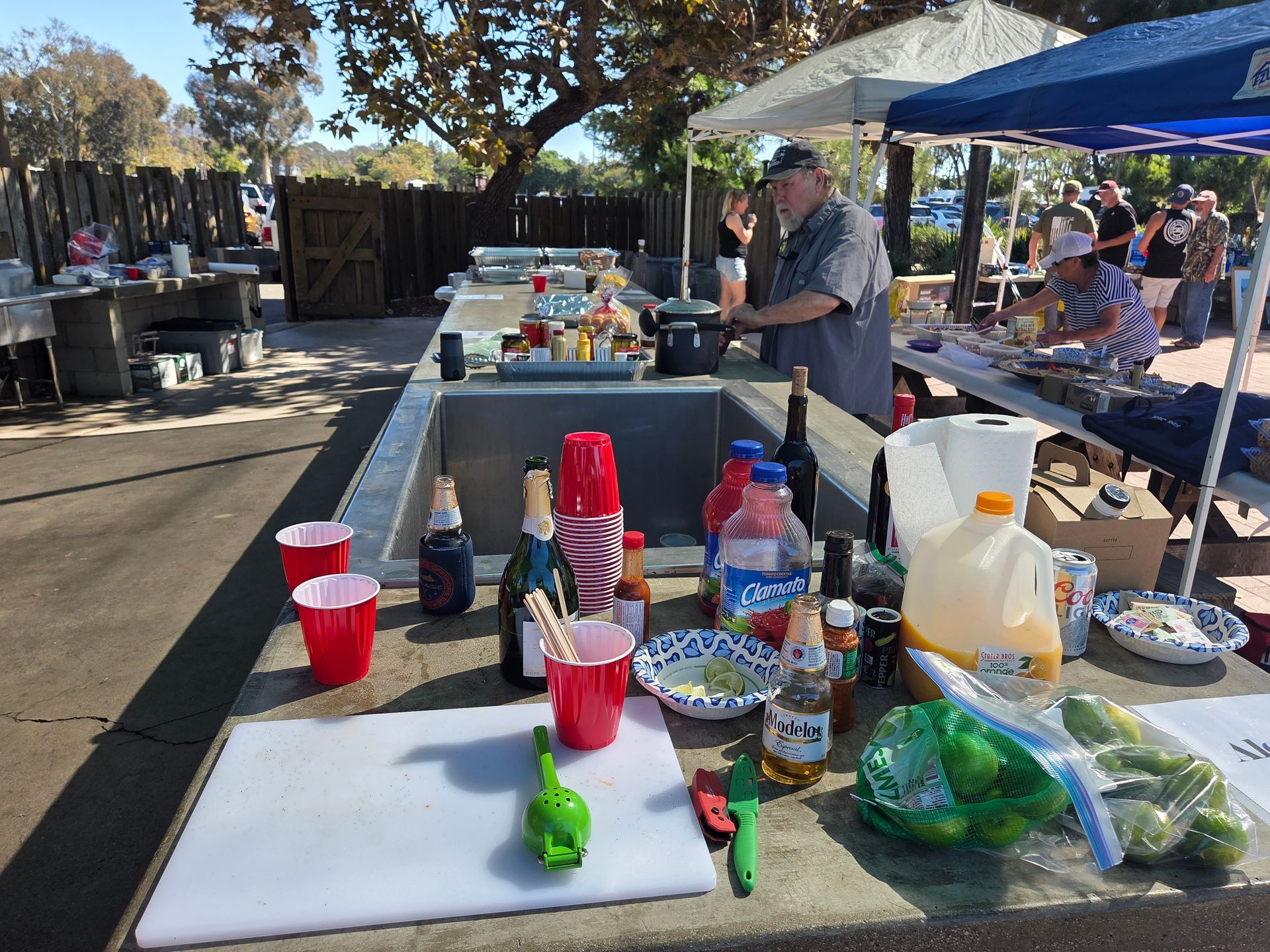 Outdoor food preparation area with various condiments, drinks, and ingredients on a concrete counter; people in the background.
