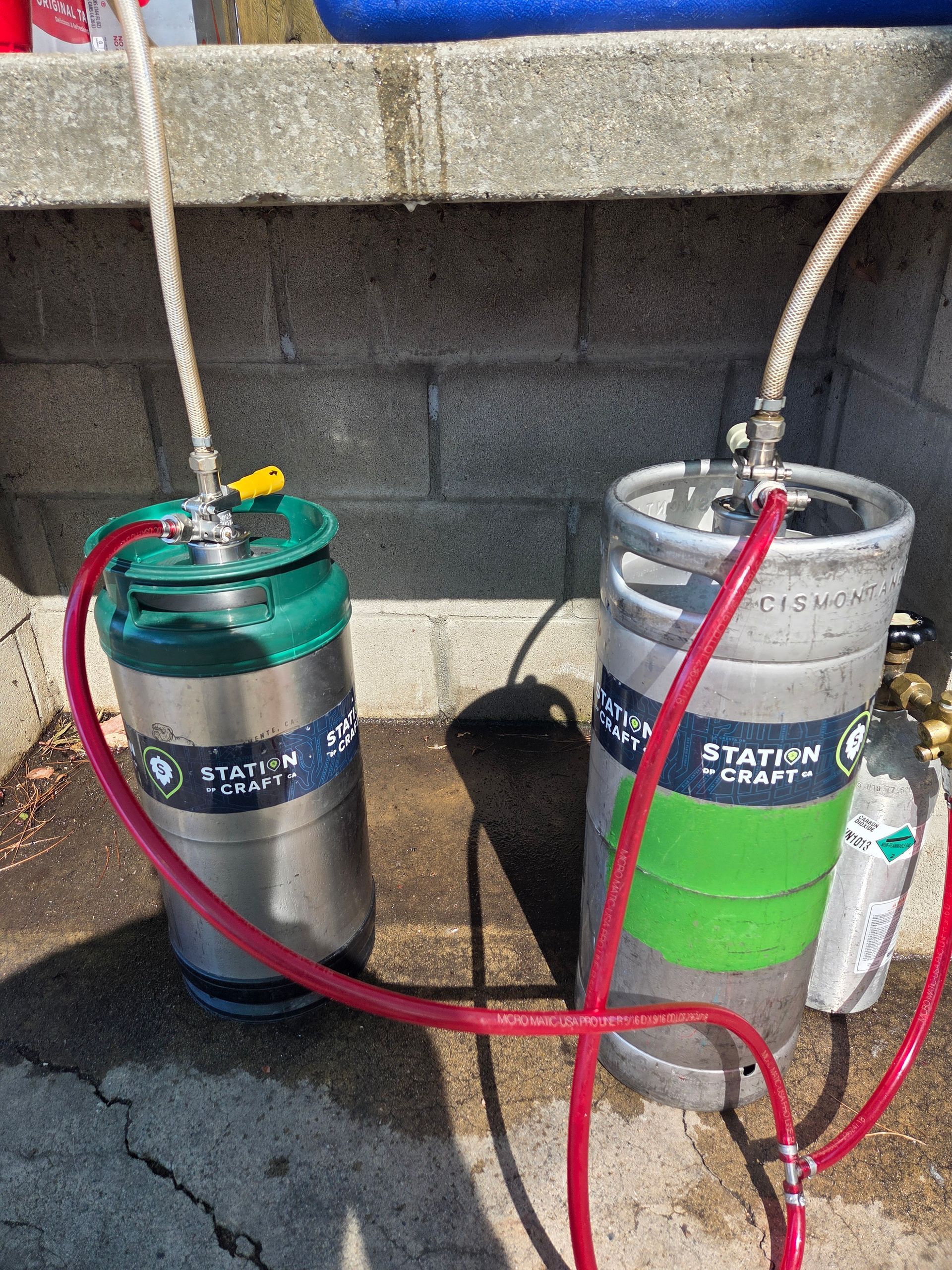 Two beer kegs with red hoses and dispensing equipment, outdoors against a brick wall.