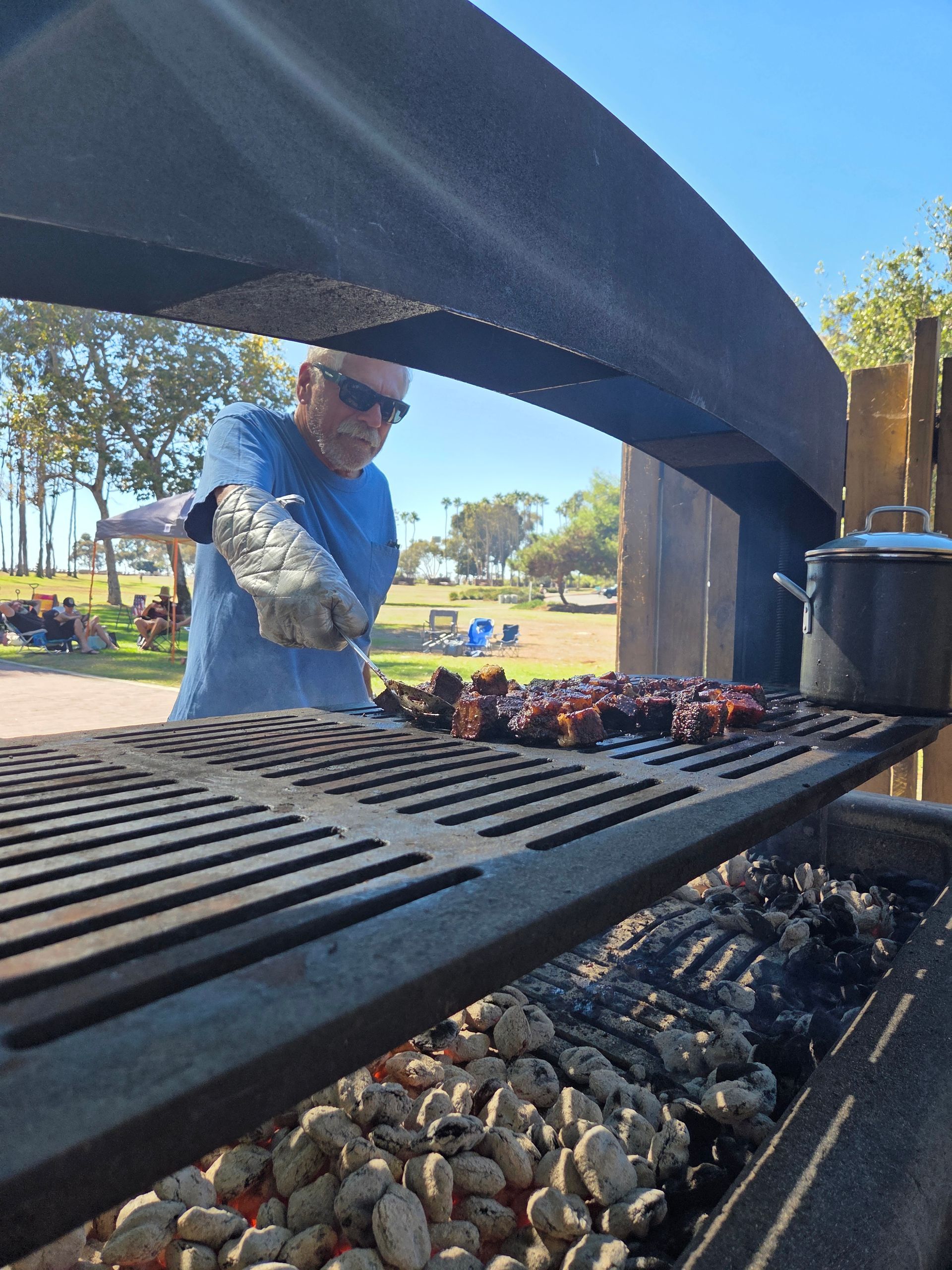 Man grilling ribs on a large charcoal grill in an outdoor park setting.