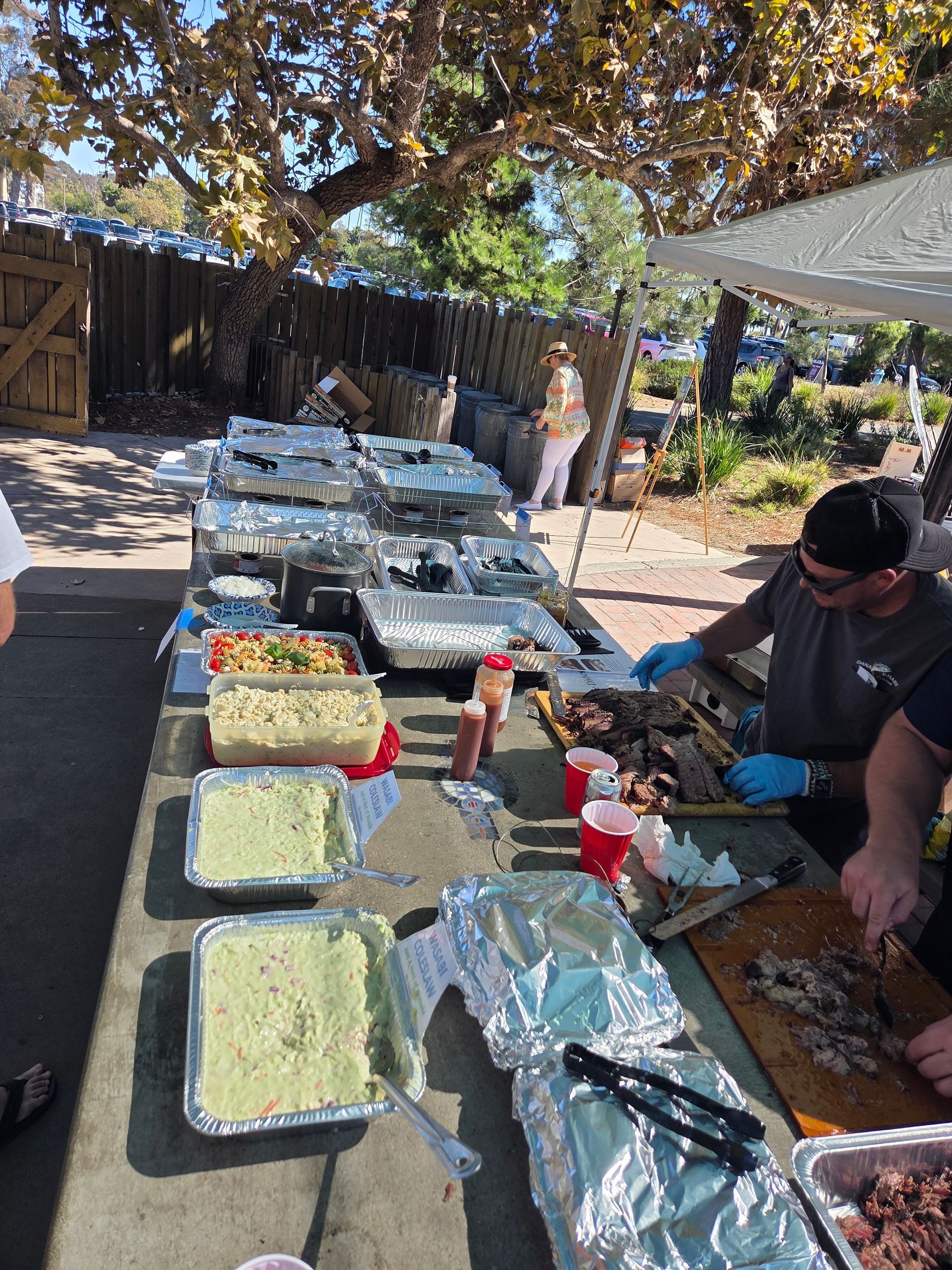 Outdoor barbecue with food on tables, people grilling and serving, sunny day.
