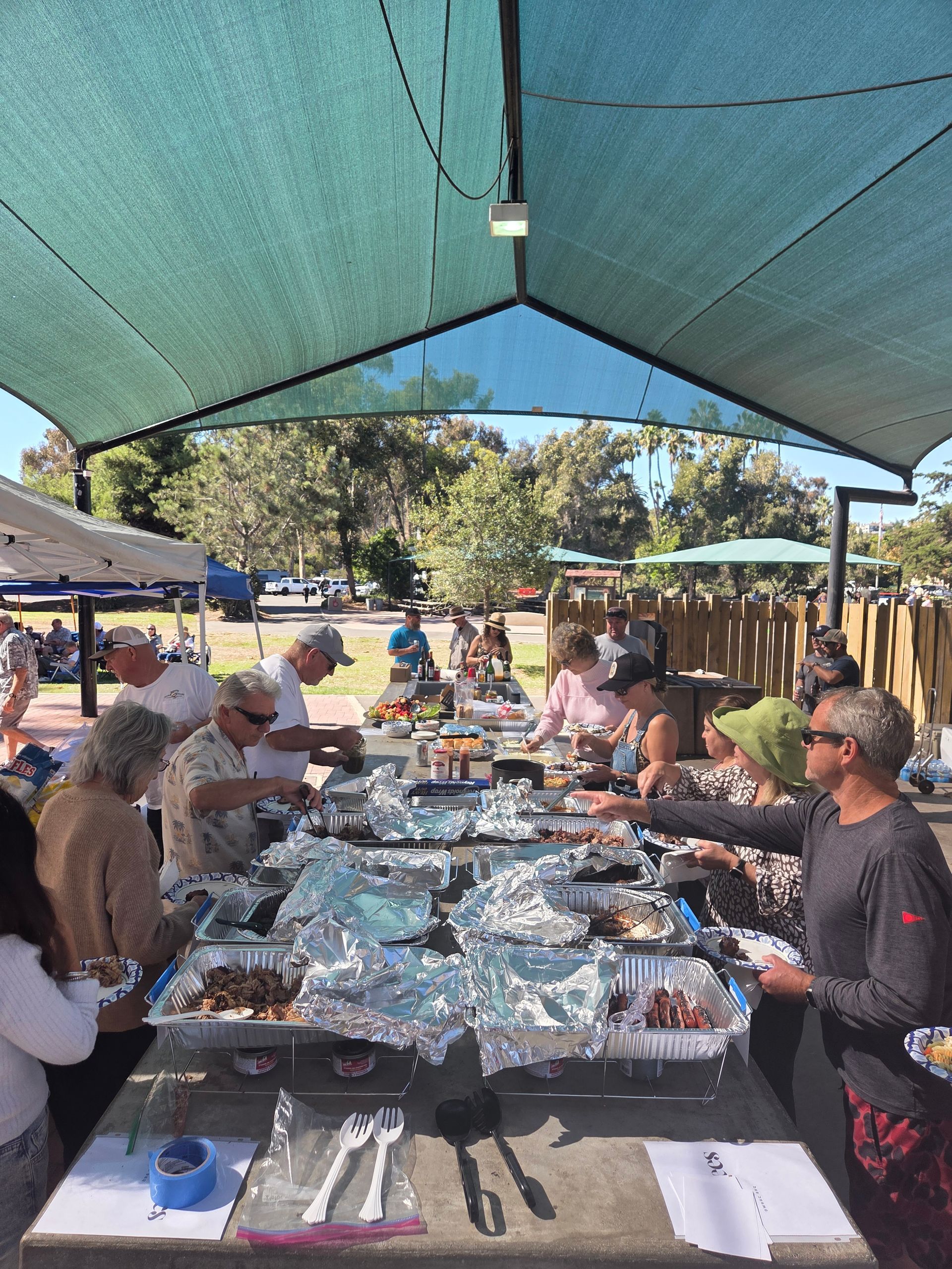 People serving food from foil-covered trays on a picnic table under a shaded structure.