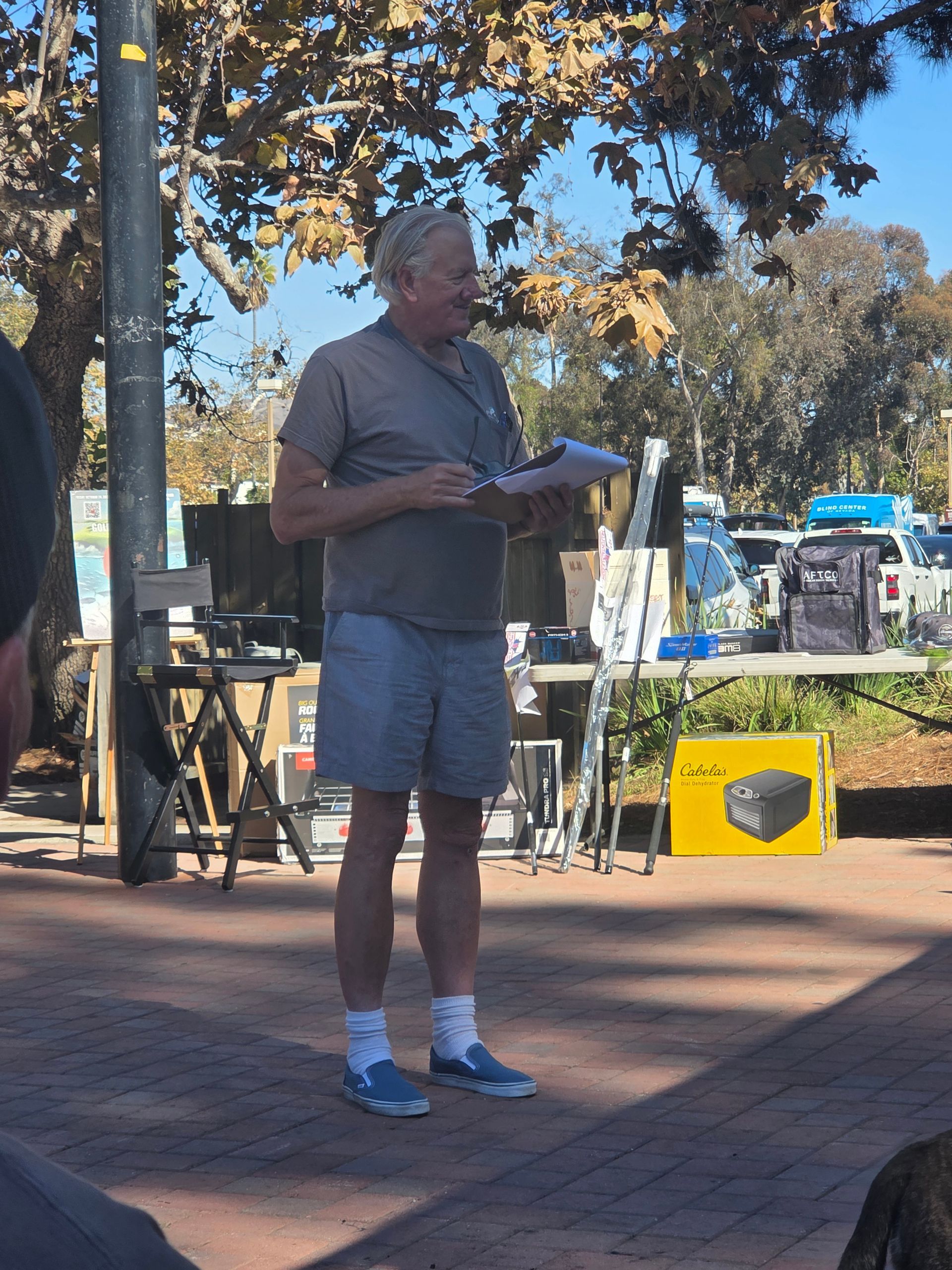 Man in shorts speaks into an outdoor crowd, holding papers.  Tables, products, and a sunny background.
