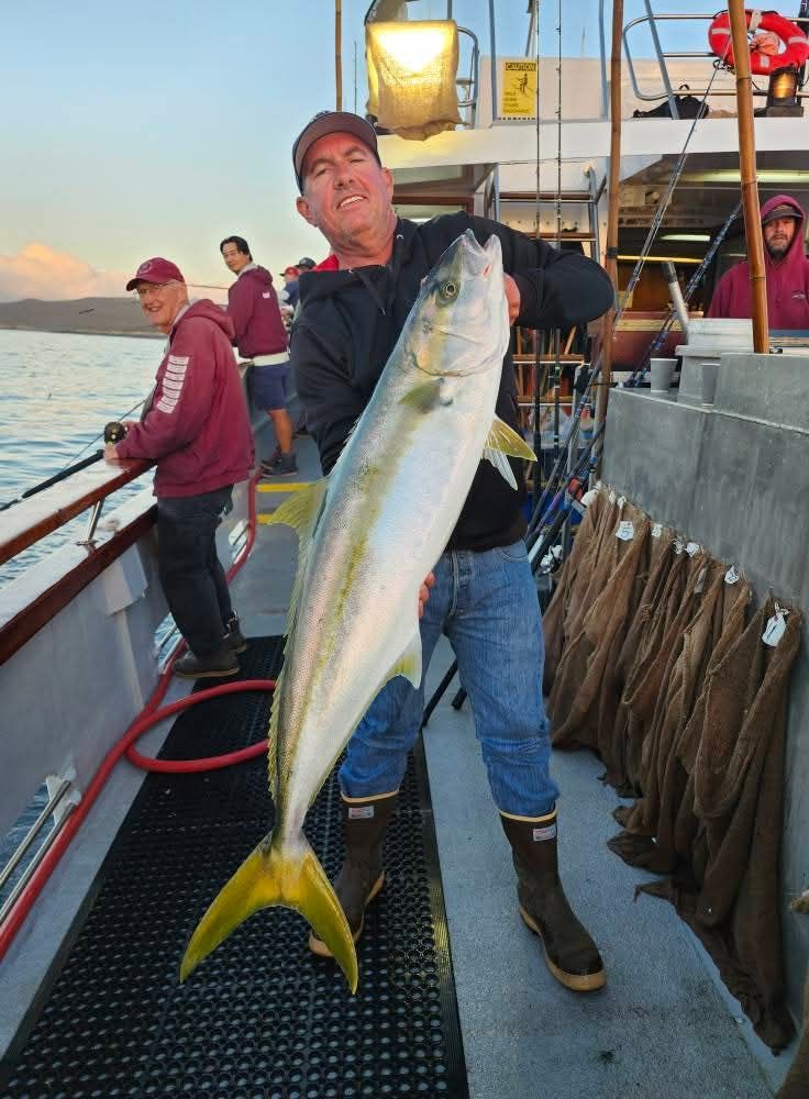 Man holding a large yellowtail fish on a boat, with other fishermen in the background.