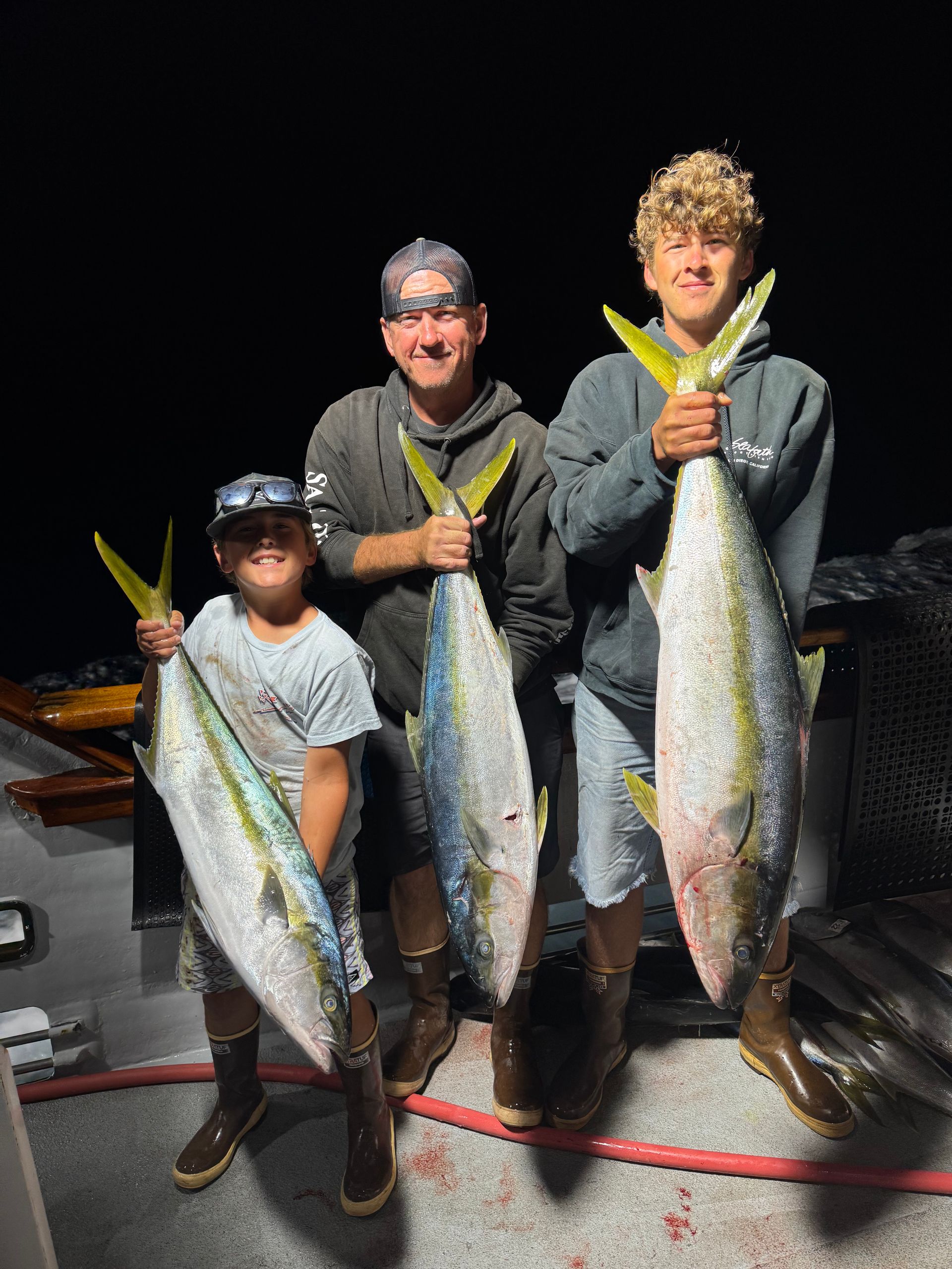 Three people holding large fish on a boat at night, smiling after a successful catch