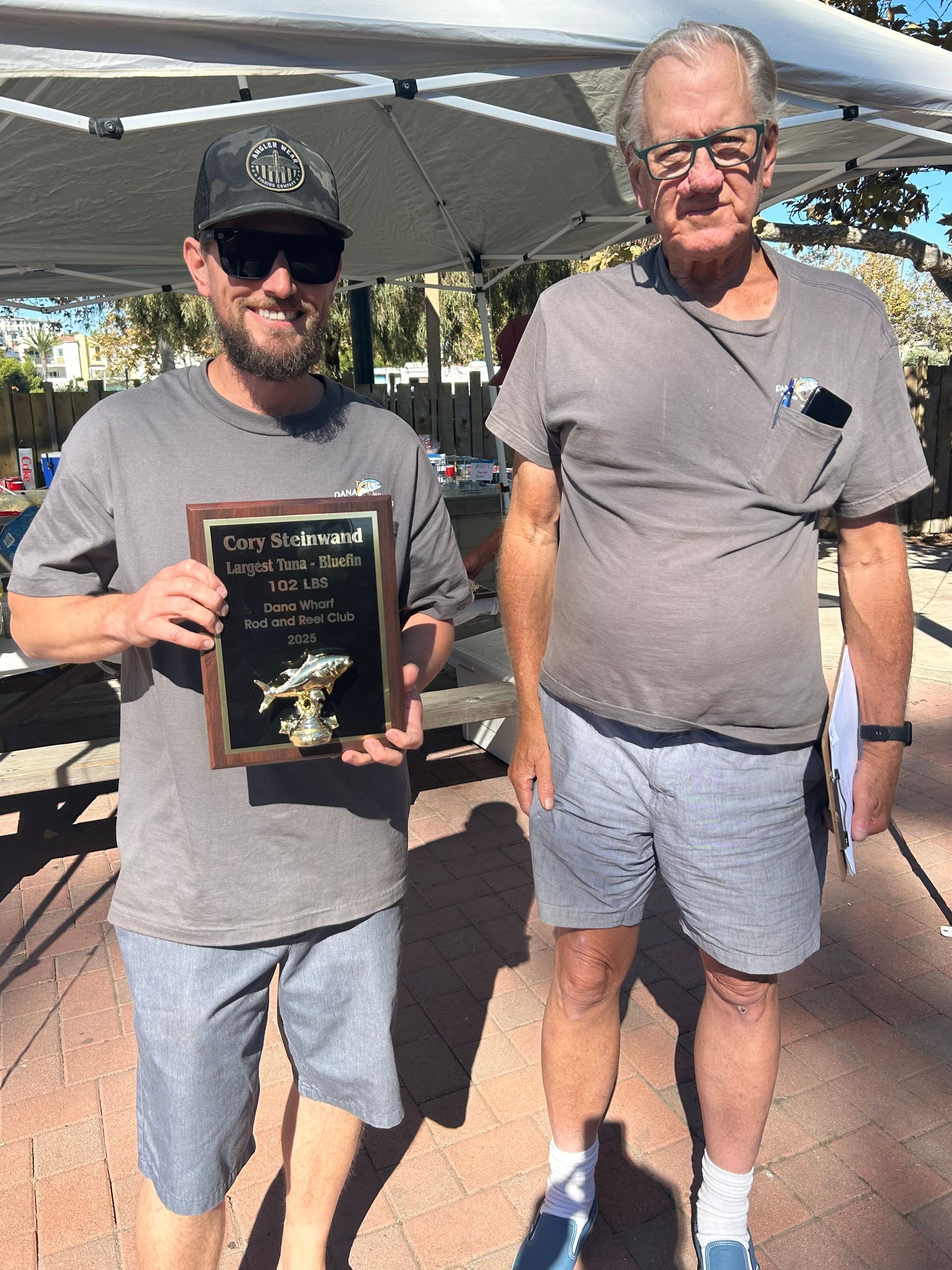 Man holding trophy with older man; outdoor setting with shade.