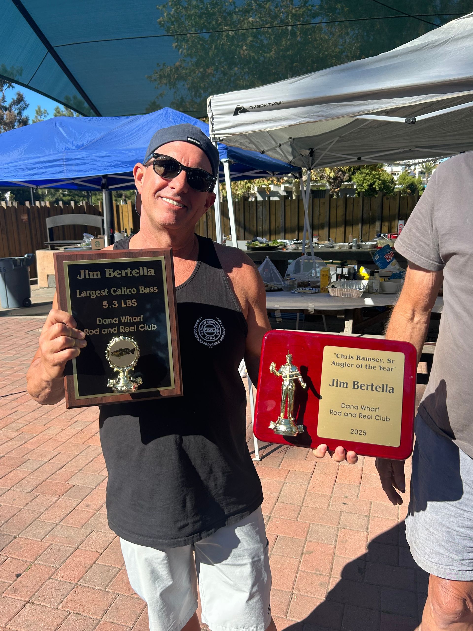 Man holding two trophies, smiling, outside with tents in the background.
