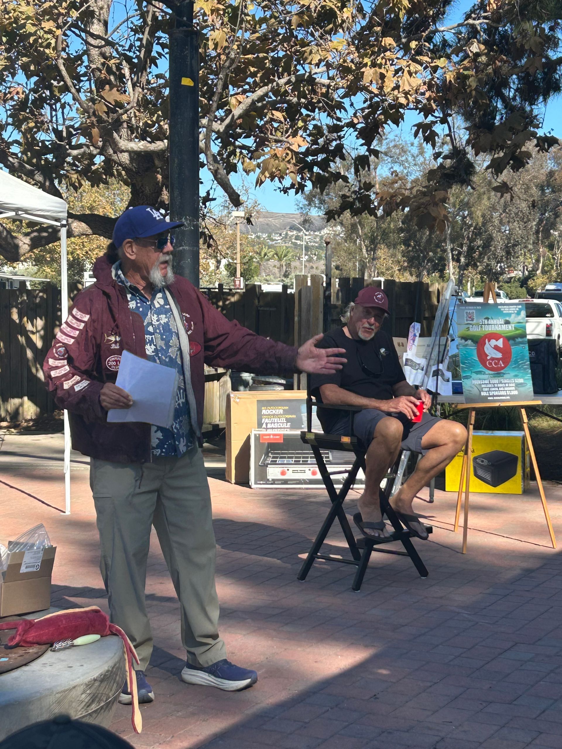 Two men outdoors: one speaking, gesturing; the other seated. Setting: sunny, public space with display.