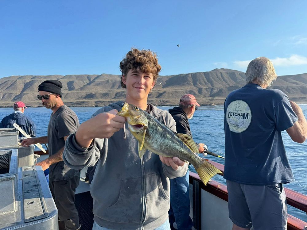 Two men on a boat hold up a large yellowtail fish with a mountainous background.