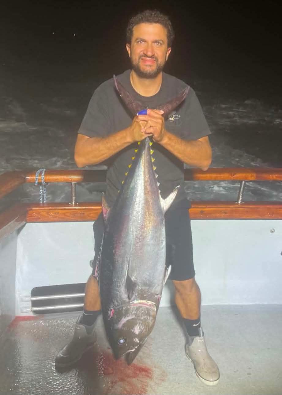 Man on boat holds up a large, silvery fish. Ocean in background.