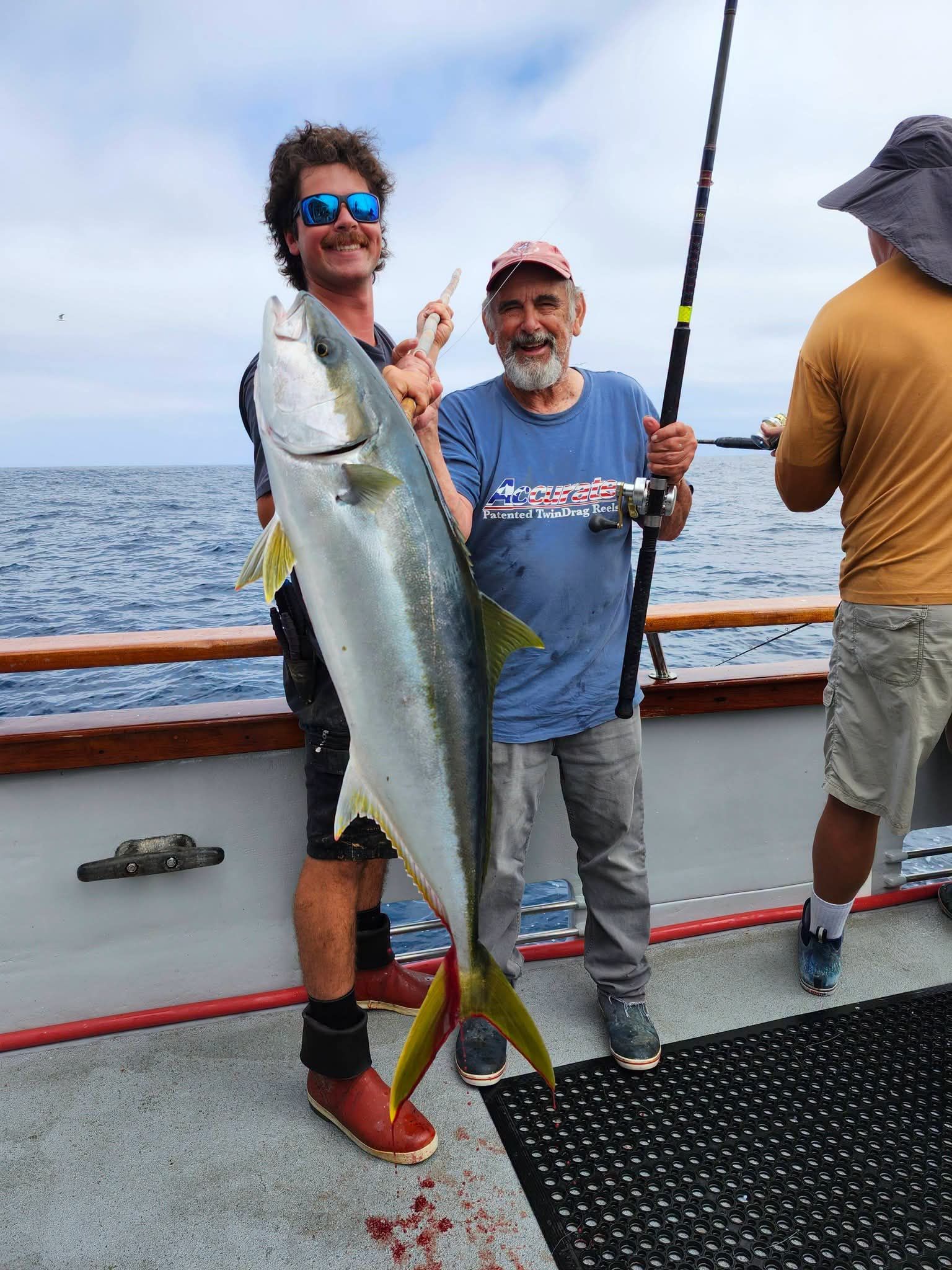 Boy holding a large yellowtail fish on a boat, giving a thumbs up. Ocean background.
