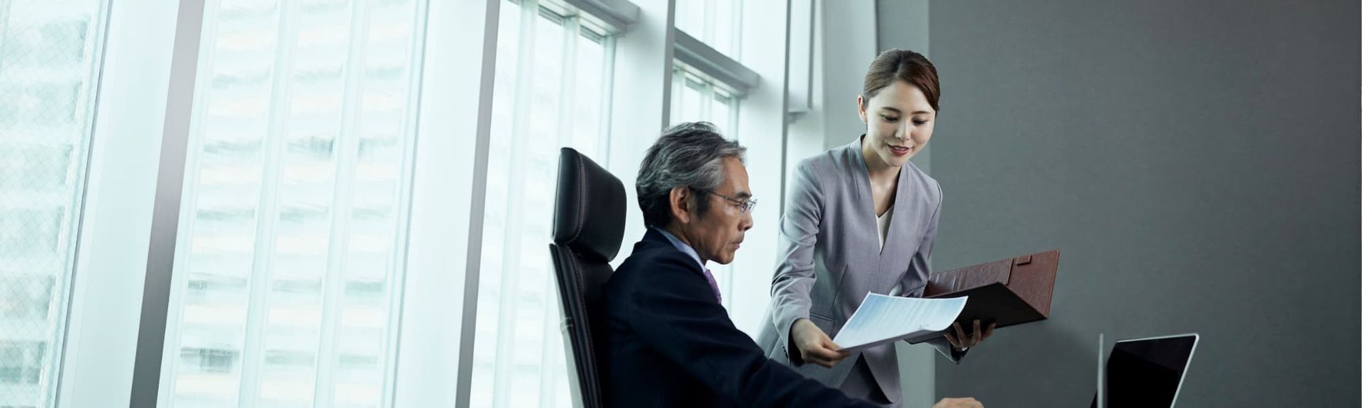 A person presents documents to an associate seated at a desk in a bright, modern office with floor-to-ceiling windows.
