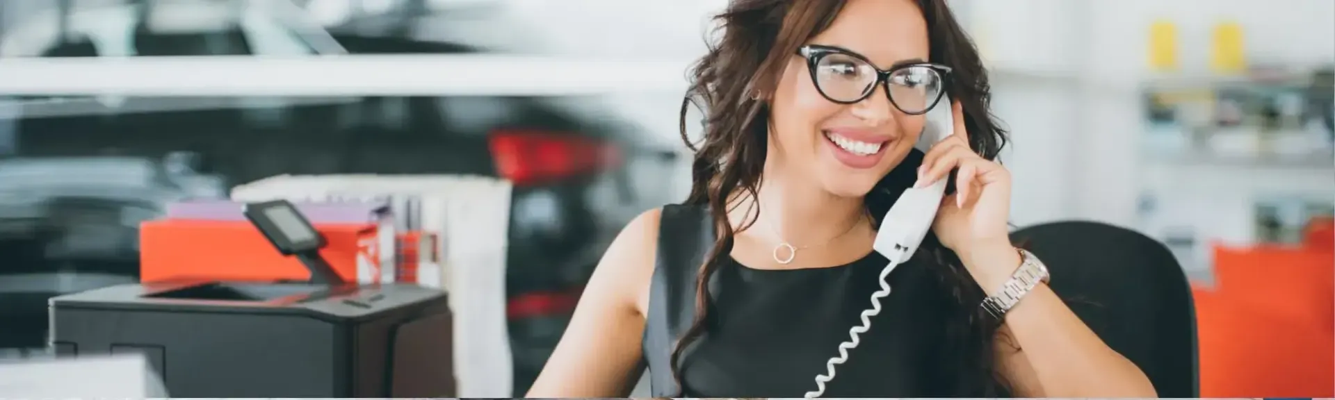 A smiling person in a dark sleeveless top and glasses talks on a desk phone in a car dealership office.