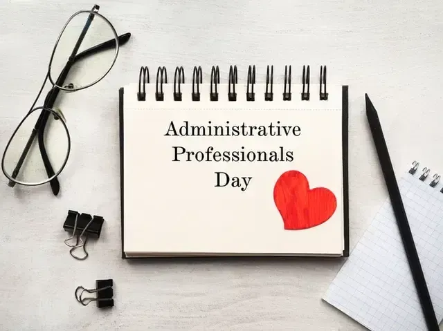 Notebook with “Administrative Professionals Day” text and a red heart, beside glasses, pen, and paper on a desk.