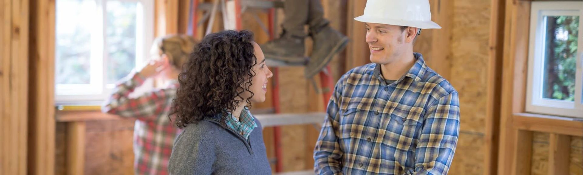 Two people discuss a home construction project in an unfinished room with wooden framing and installed windows.