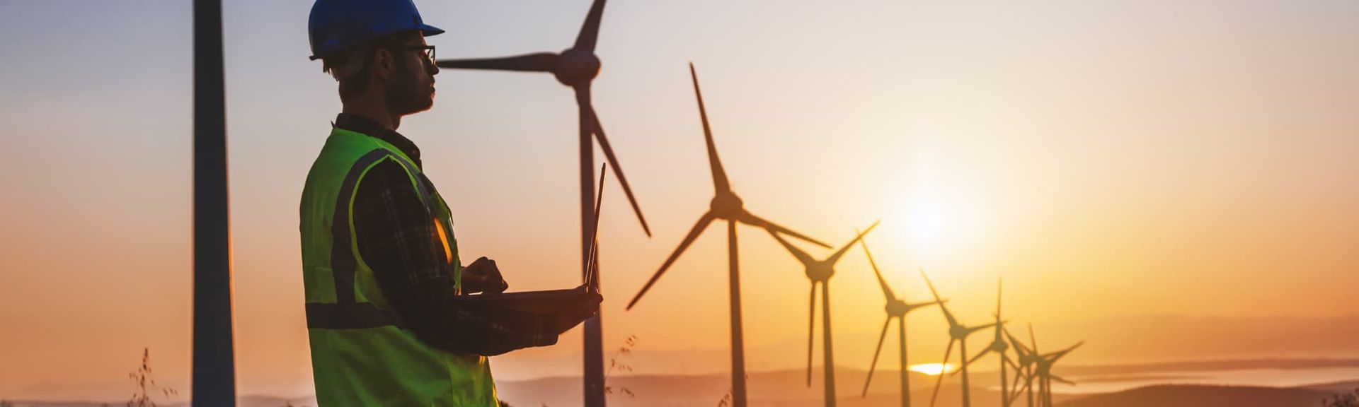 Worker in safety gear beside wind turbines at sunset, with a warm orange sky.