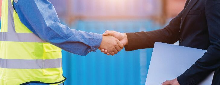 A person in a high-visibility vest shakes hands with someone in a suit holding a folder, with shipping containers behind.
