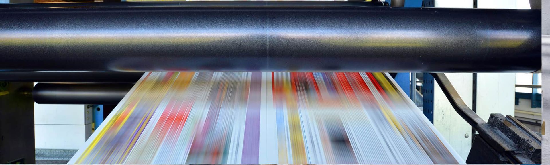 A colorful, patterned sheet of material moving horizontally under a large, dark industrial roller in a factory setting.