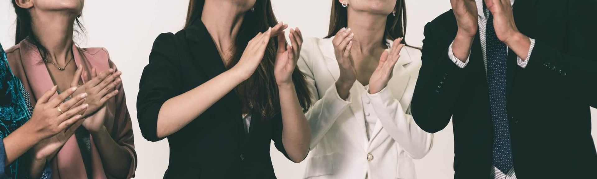 Four individuals in professional attire clapping their hands while looking upward against a plain background.