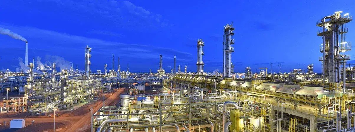 An industrial oil refinery with tall distillation towers and illuminated pipes against a dark blue twilight sky.