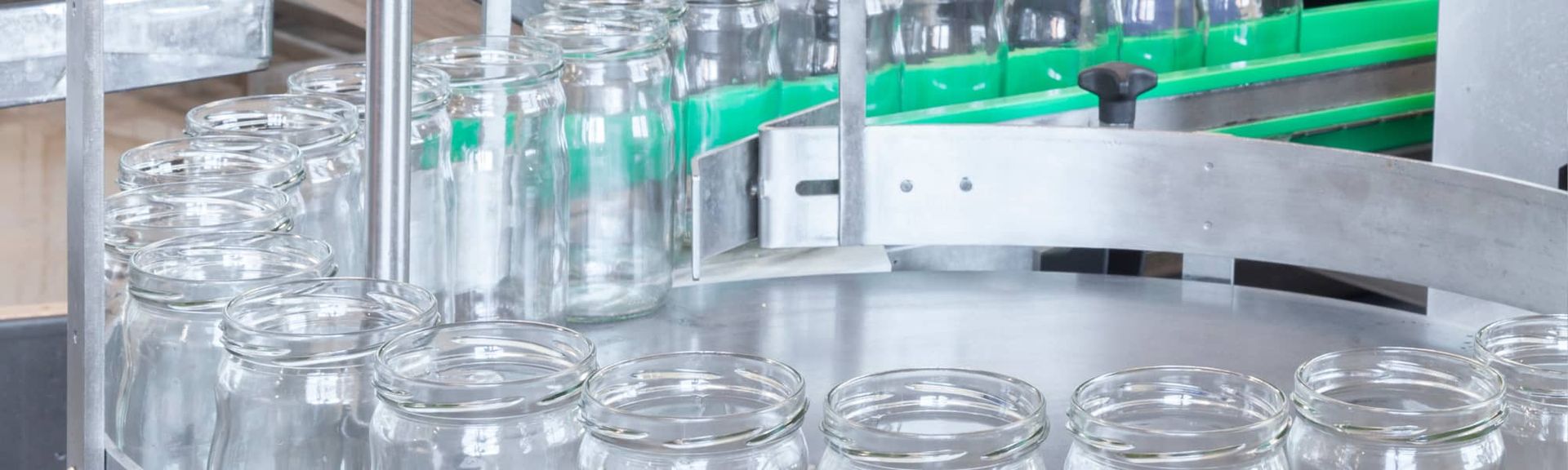 Clear glass jars move along a metallic industrial conveyor belt in a manufacturing facility.