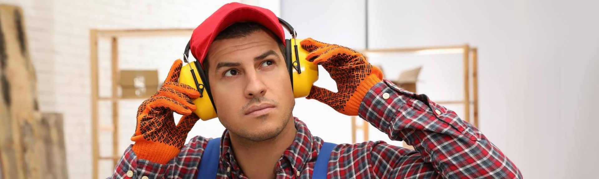A worker in a red cap, plaid shirt, and gloves adjusts yellow ear protection in a workshop.
