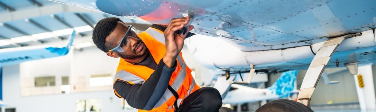 Technician in orange safety vest inspecting wiring inside an aircraft cabin ceiling panel