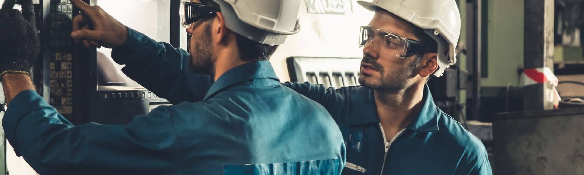Two workers in hard hats and safety goggles standing in a factory, one pointing at machinery while the other watches.