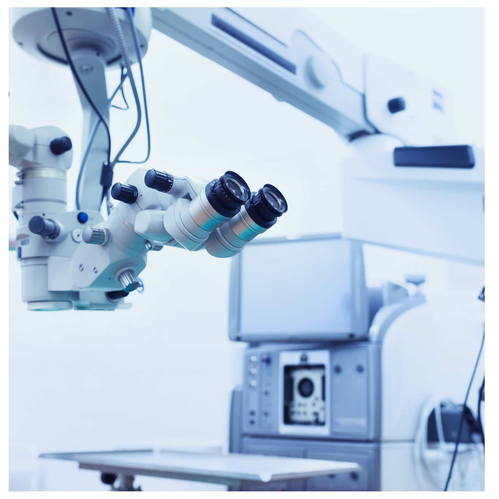 Surgical microscope in a bright operating room, with a monitor and robotic arms visible in the background