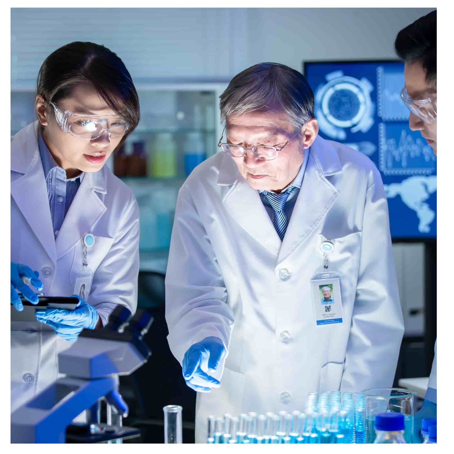 Scientists in white lab coats examine samples at a microscope in a blue-lit laboratory.