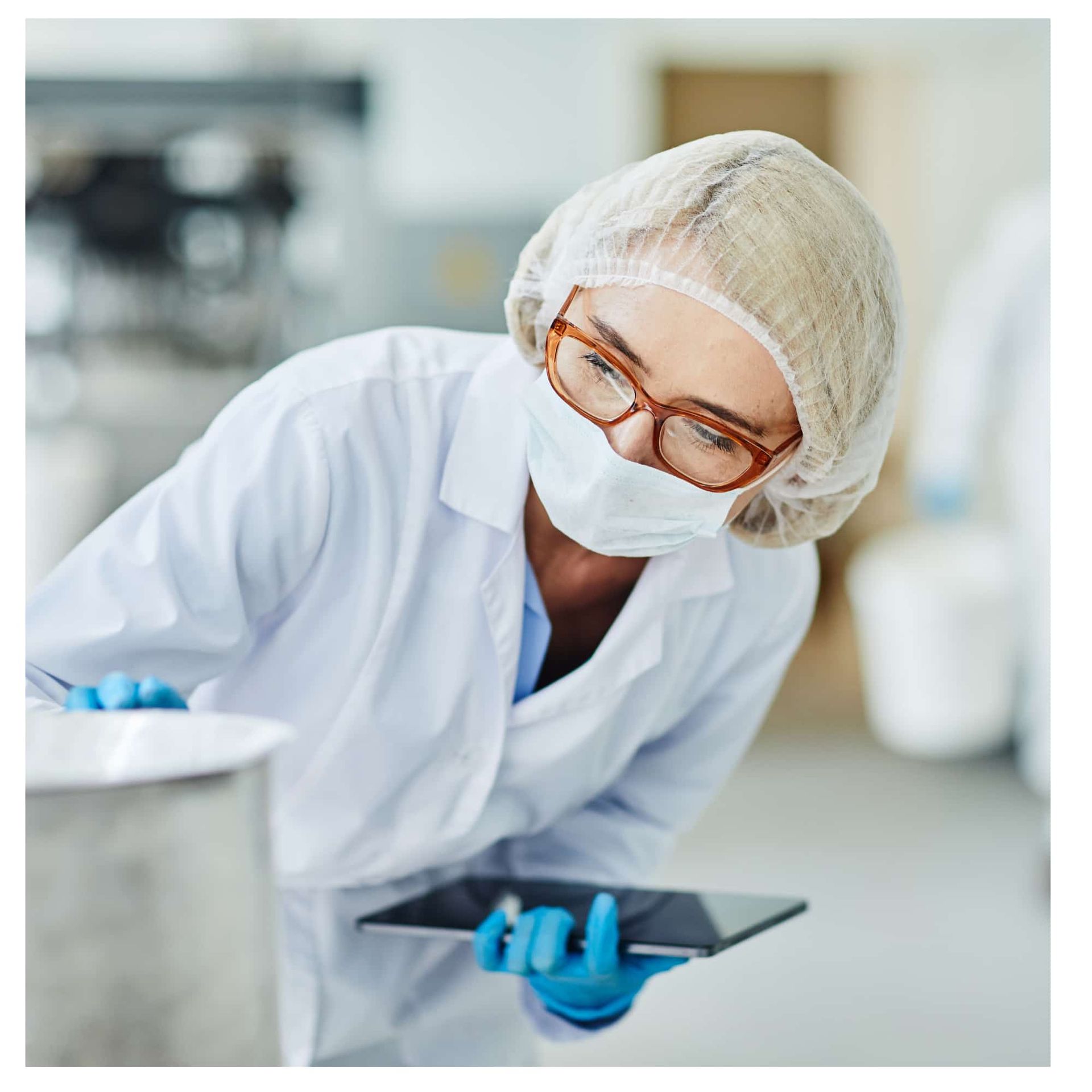 Lab technician in white coat, hairnet, and mask using a tablet in a bright lab
