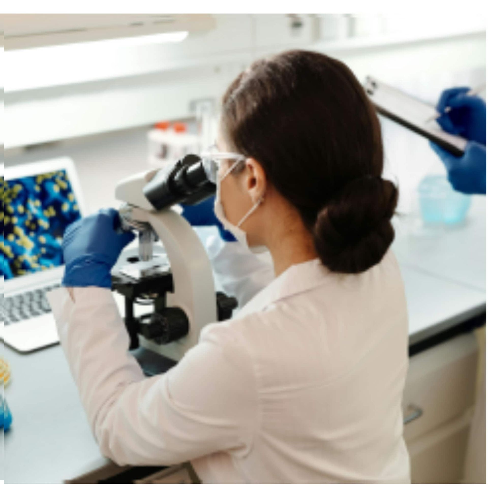 Scientist in lab coat using a microscope in a laboratory, with blue-gloved hands and a computer screen nearby