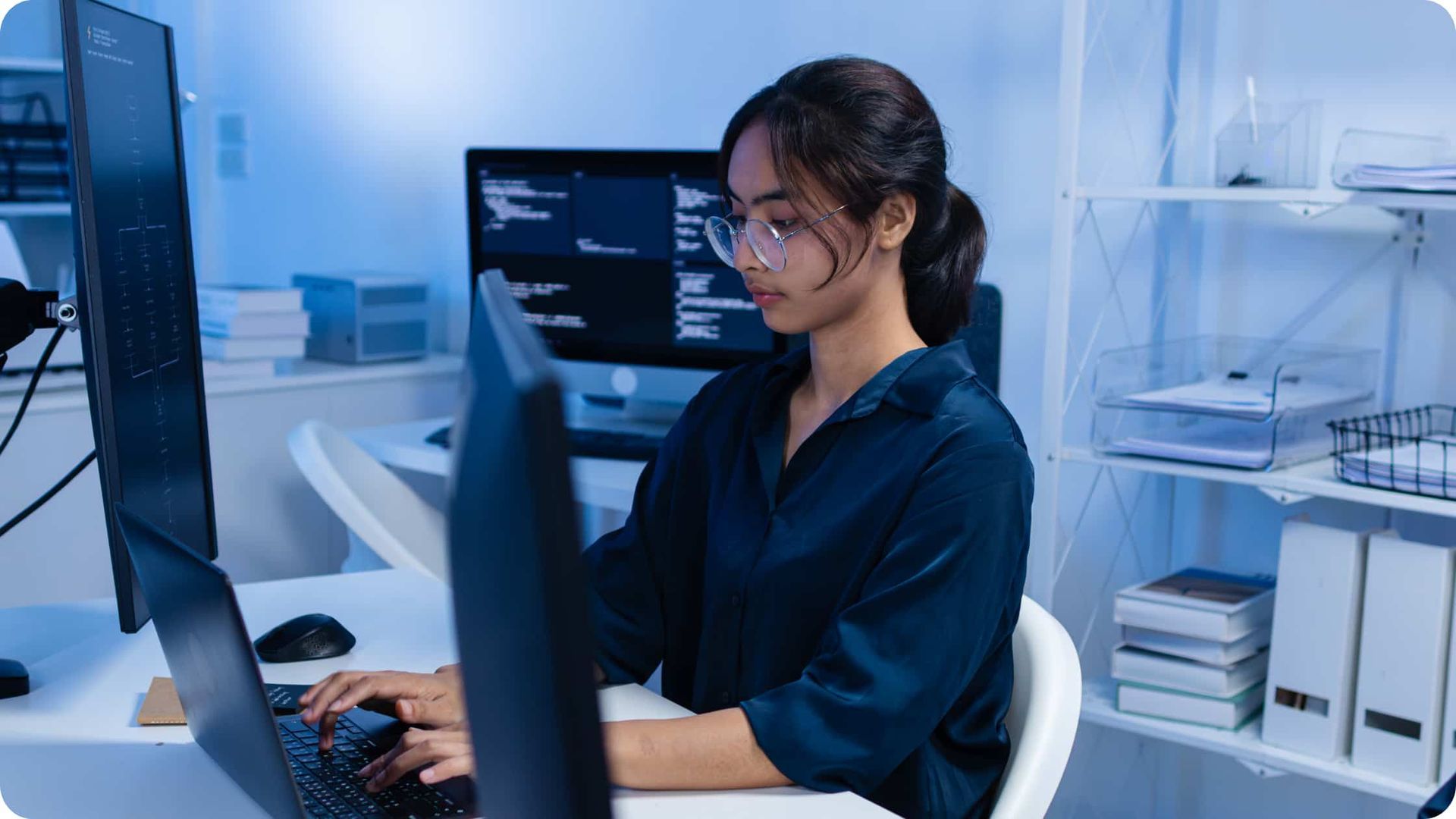 Person working at a desk with dual monitors in a blue-lit office, typing on a keyboard.