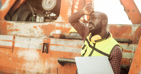 Worker in a yellow vest and hard hat stands by an orange industrial vehicle, shading his eyes.