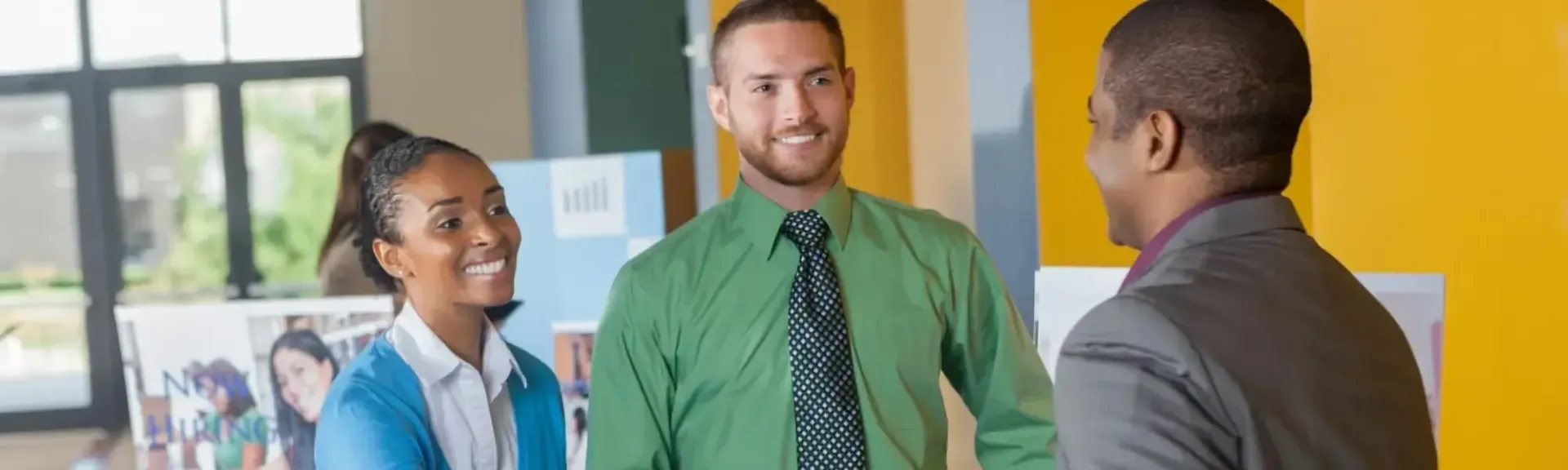 Three colleagues in business attire converse and smile in a bright, modern office with displays in the background.