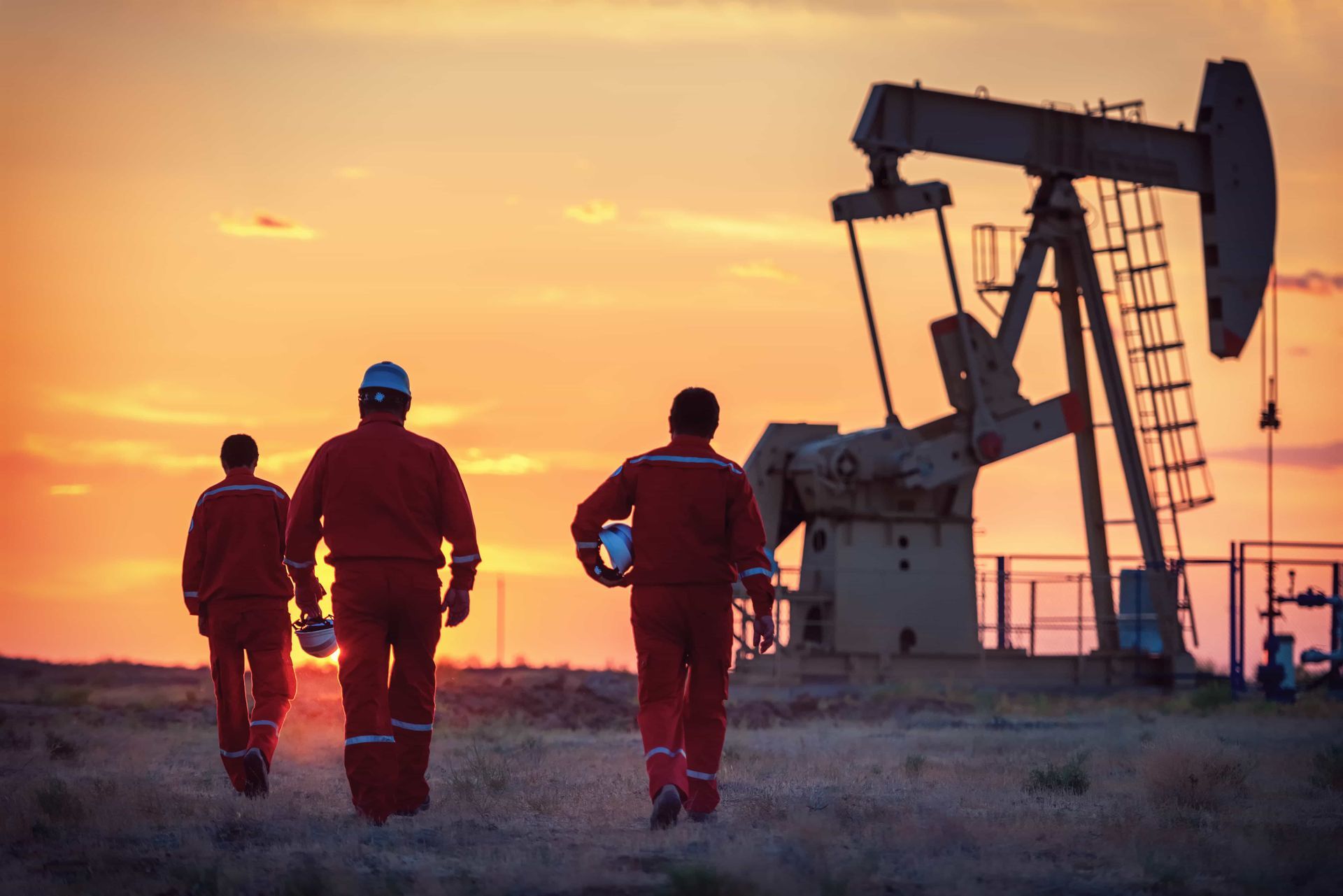 Three workers in orange coveralls walk toward an oil pumpjack at sunset.