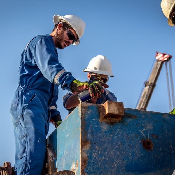 Two construction workers in blue coveralls and hard hats working on a metal structure outdoors.