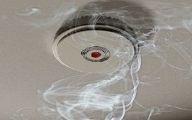 A smoke detector mounted on a ceiling with wisps of white smoke rising toward it.