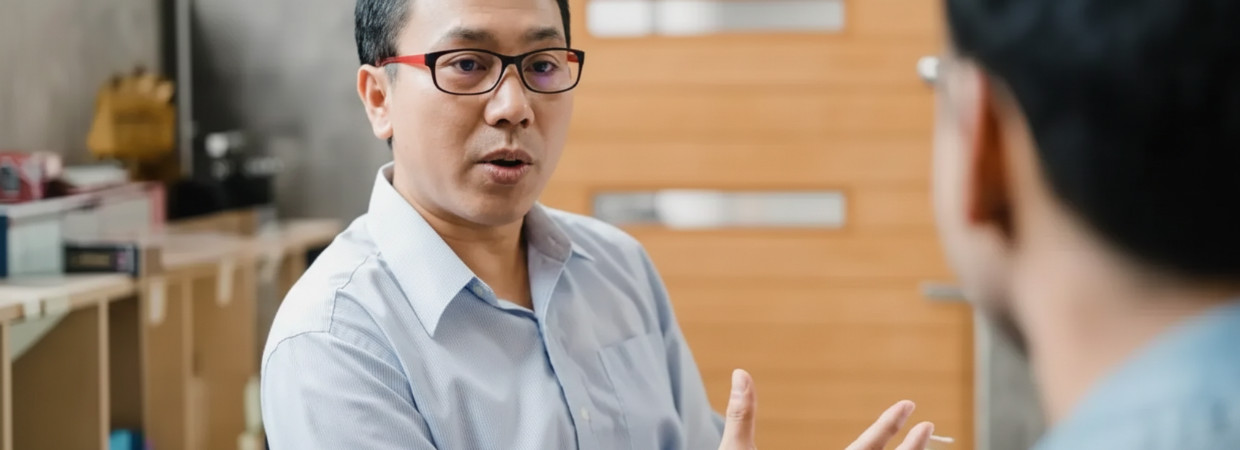 A person in a light blue shirt wearing glasses speaks with someone off-camera in an office setting.