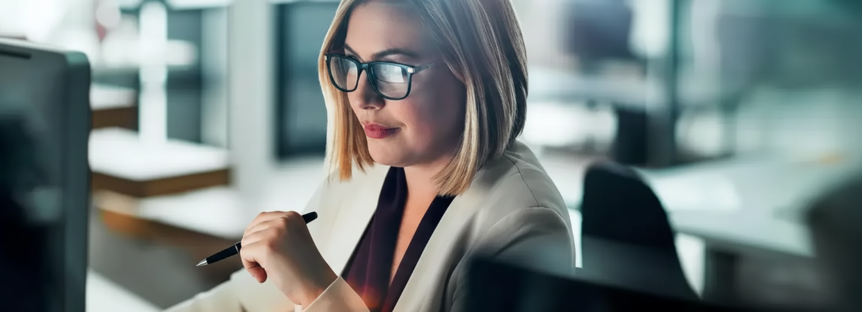 A person wearing glasses holds a pen while looking intently at a computer screen in a professional office setting.