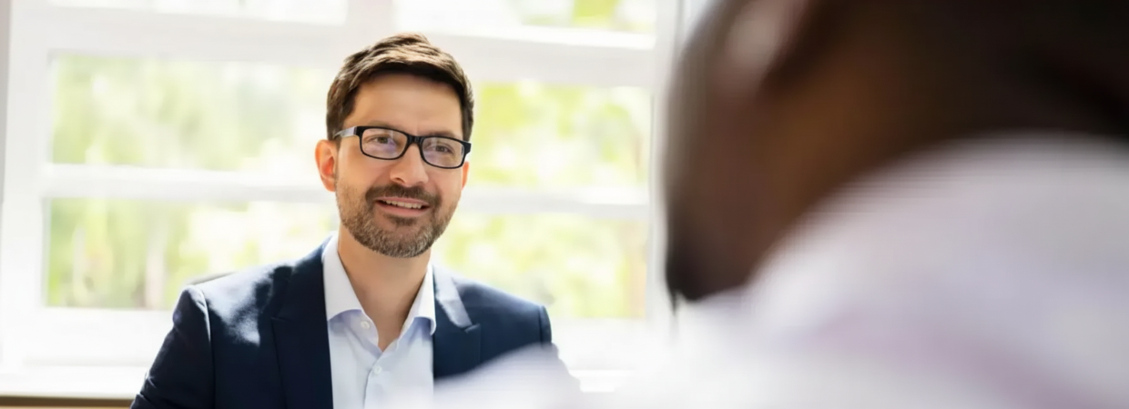 A person in a business suit and glasses smiles while engaged in a conversation with someone off-camera.