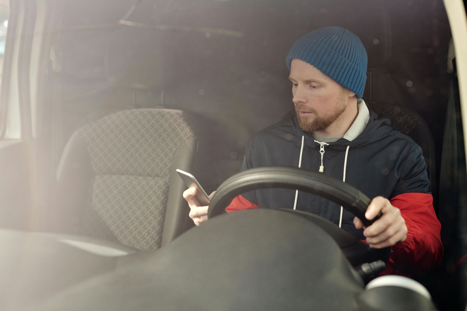 A person wearing a blue beanie and dark hoodie with red sleeves sits in a vehicle driver's seat, looking at a smartphone.