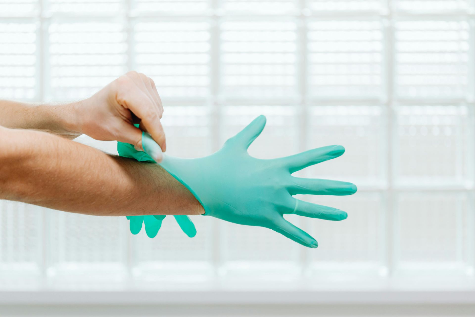 A pair of hands pulls on a teal medical glove against a background of glass block windows.