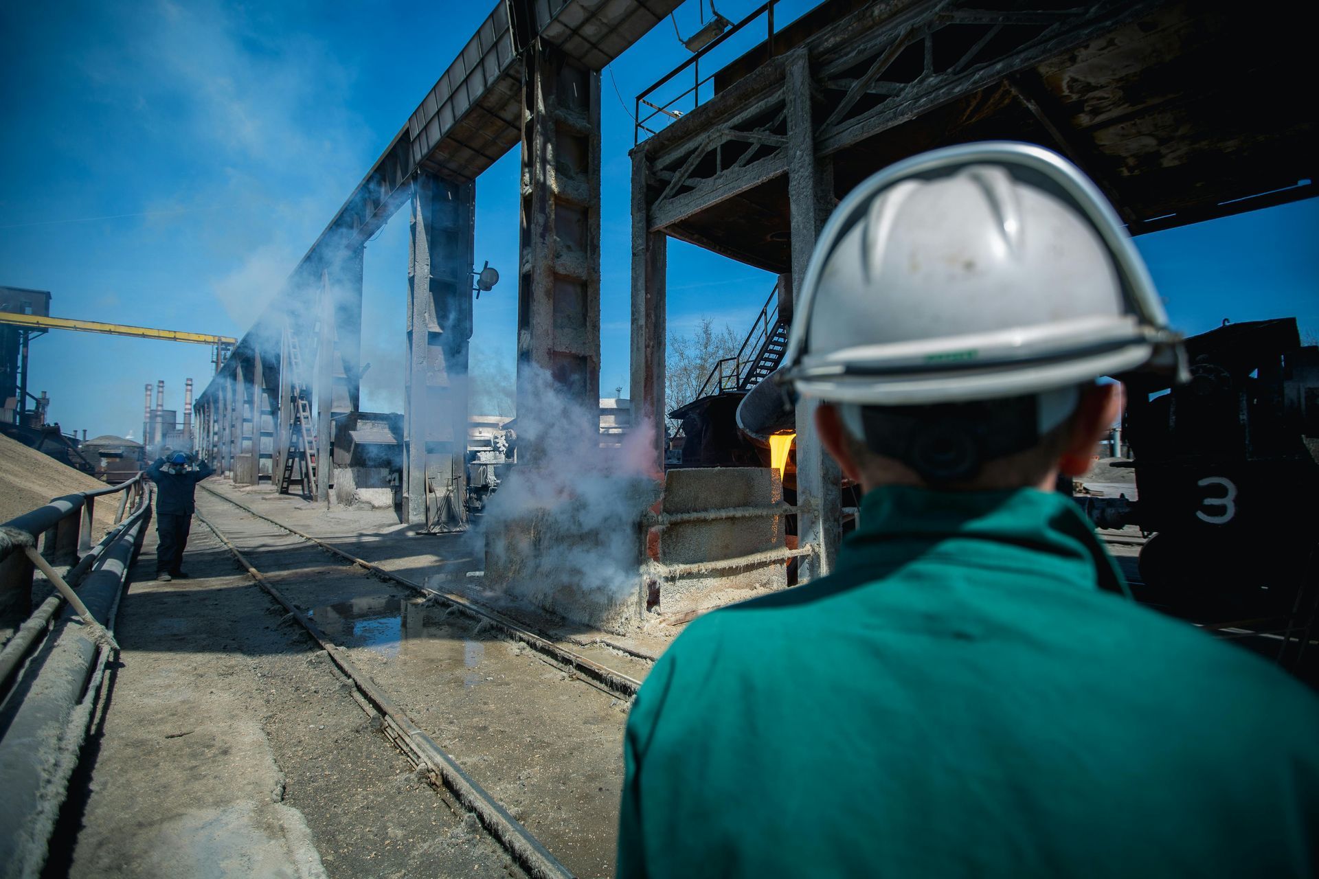 A worker in a green jacket and white hard hat overlooks a smoking industrial site with a large, elevated rail structure.
